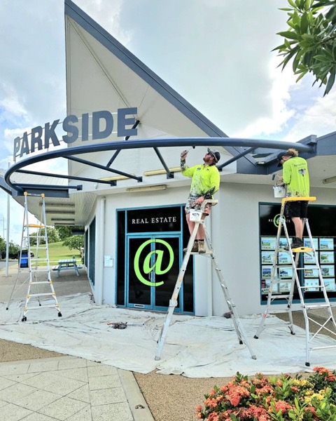Two painters in branded shirts on ladders coating the curved awning of a shopfront