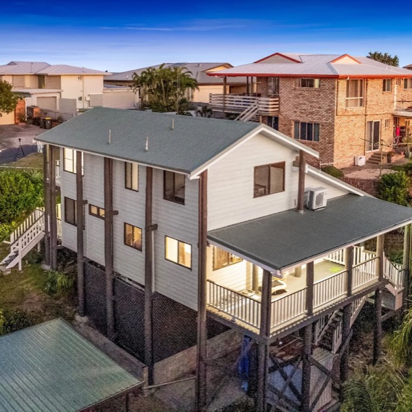 A highset home with white cladding and a dark roof on a sloping block